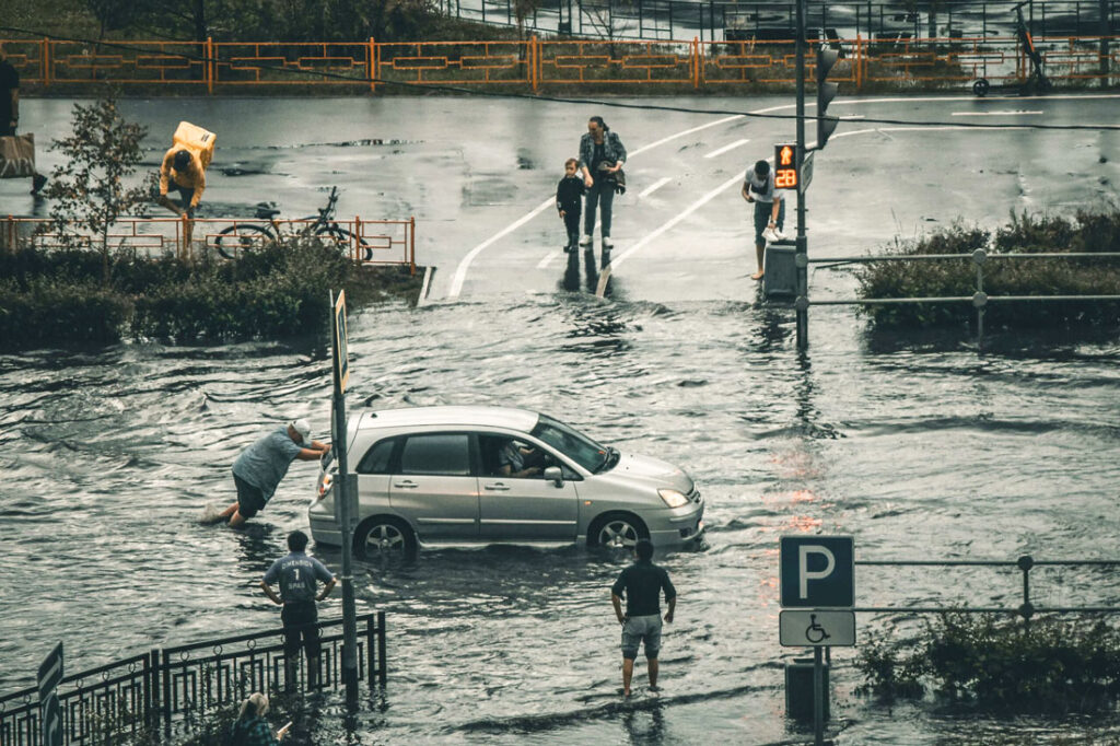 Eine Straße steht unter Wasser. Ein Mann versucht sein Auto von der überfluteten Straße zu schieben.
